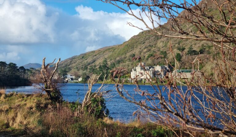 A scenic view of Kylemore Abbey in Ireland, nestled at the foot of a forested hill and reflected in a calm lake. Leafless branches in the foreground frame the historic stone building, set against a backdrop of rolling mountains and a bright blue sky with scattered clouds.