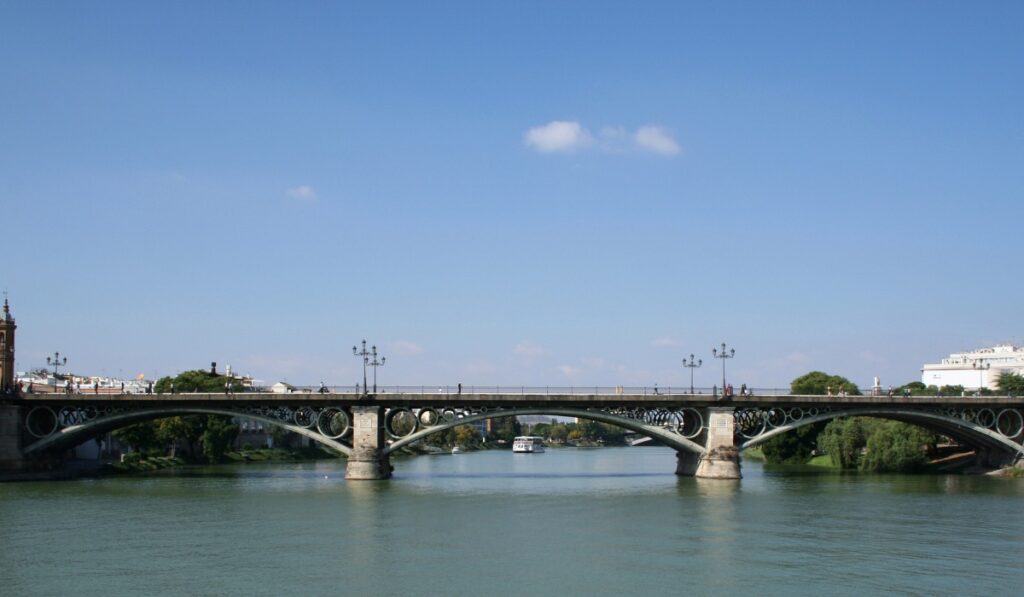 The Isabel II Bridge, also known as the Triana Bridge, stretches across the Guadalquivir River in Seville, Spain. The 19th-century cast iron bridge features elegant arches and vintage street lamps, with a boat passing beneath and pedestrians strolling along the top.