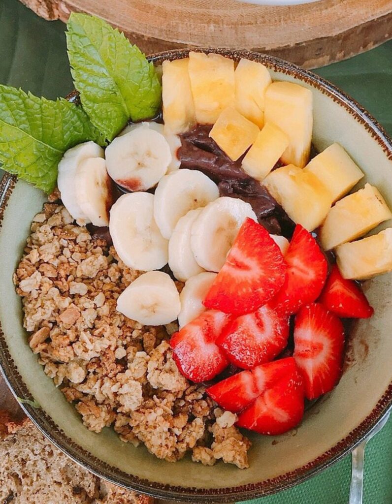 A colorful acai bowl topped with sliced bananas, chopped pineapple, halved strawberries, granola, and fresh mint leaves. The toppings are neatly arranged in sections over a dark acai base, served in a ceramic bowl on a green tablecloth.