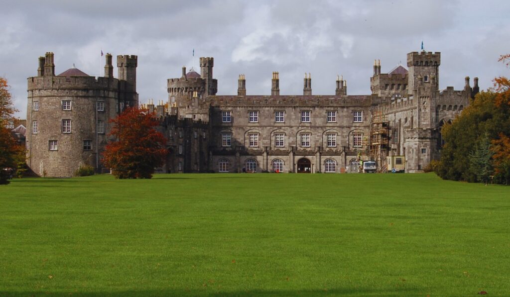 Kilkenny Castle in Ireland, a grand stone fortress with towers and battlements, stands against a cloudy sky and is surrounded by vibrant green lawns. Autumn trees with golden and rust-colored leaves frame the scene, adding warmth to the historic view.