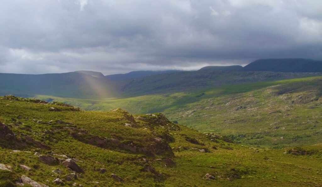 A dramatic view of Killarney National Park in Ireland, with rolling green hills, rocky outcrops, and a faint beam of sunlight breaking through thick clouds onto the valley floor. The scene captures the wild, moody beauty of the Irish countryside.