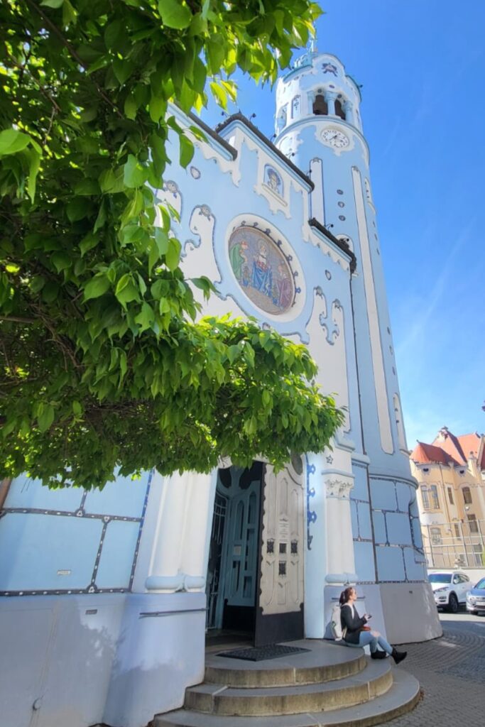 Me sitting on the steps outside the whimsical Blue Church in Bratislava, officially the Church of St. Elizabeth, with its pastel blue Art Nouveau façade partially shaded by a leafy tree.