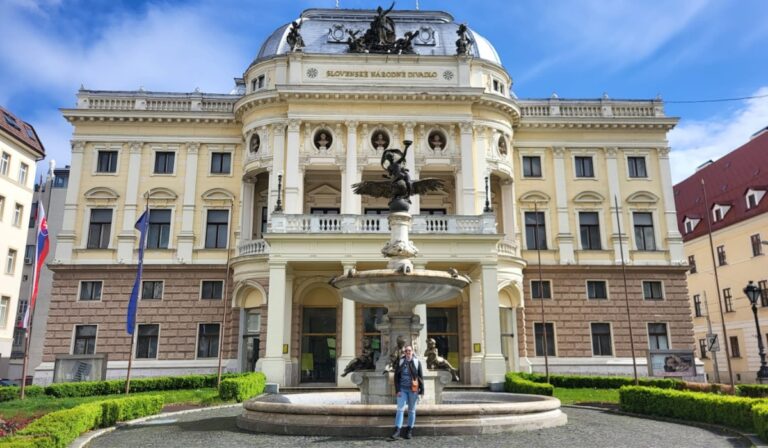 Me standing in front of the ornate fountain outside the Slovak National Theatre in Bratislava, a grand historic building with a domed roof, statues, and classical columns. The scene is framed by flags, trimmed hedges, and a clear spring sky.
