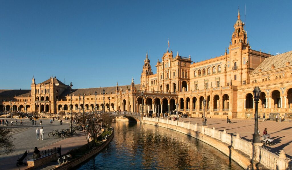 A wide-angle view of Plaza de España in Seville, Spain, showing its grand semicircular Renaissance-style building, arched bridges, central canal, and people walking along the expansive plaza under a clear blue sky.