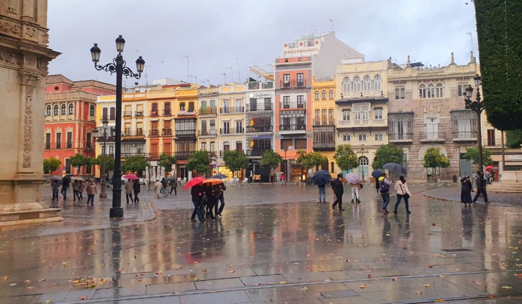 People with umbrellas walk across a rain-soaked square in Seville, Spain, surrounded by colorful historic buildings with wrought-iron balconies and orange trees. The wet pavement reflects the soft, cloudy light and vibrant facades.