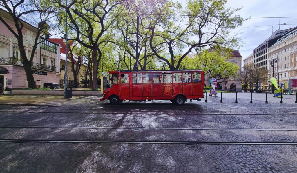 A bright red vintage-style tour bus sits parked on a cobblestone street in Bratislava, with leafy trees and historic buildings in the background. The scene includes tram tracks in the foreground and banners advertising tours near the Savoy Hotel and the Opera House.