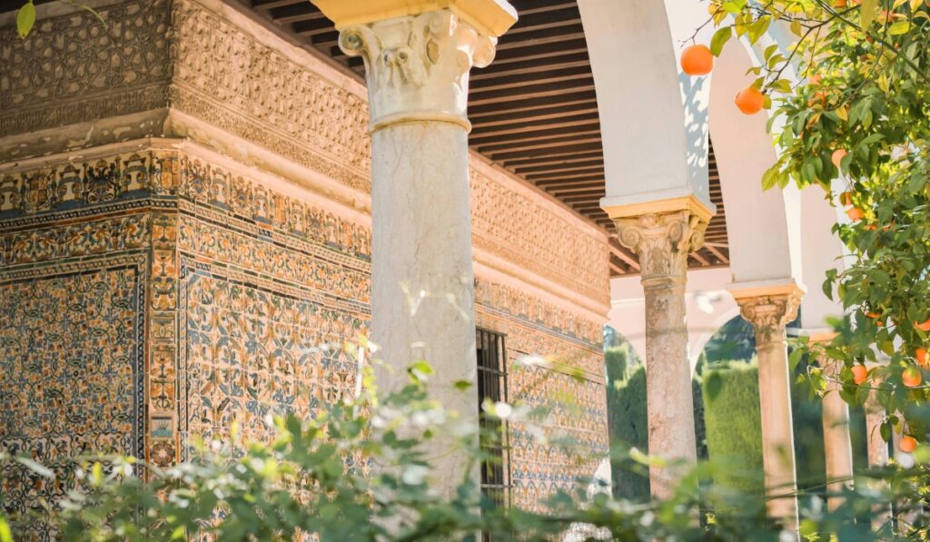 Detailed view of the Royal Alcázar of Seville featuring colorful ceramic tilework, marble columns, and white arches, with orange trees partially visible in the garden.
