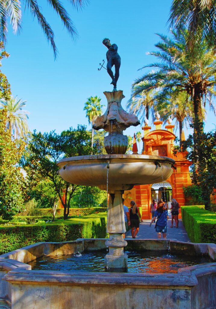 A classical fountain with a statue of Mercury in the lush gardens of the Royal Alcázar of Seville, surrounded by palm trees, trimmed hedges, and visitors walking along a sunny path.