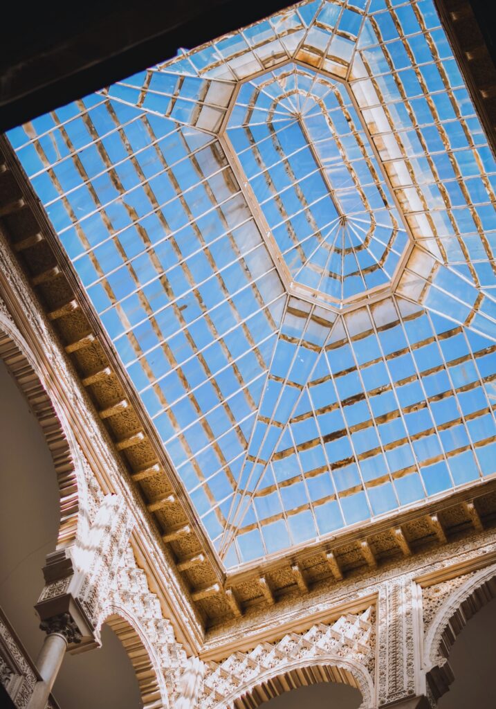Ornate glass ceiling with a geometric pattern viewed from below, framed by intricately carved arches and Moorish-style columns at the Royal Alcázar of Seville.