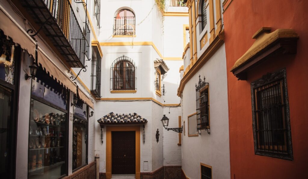 A narrow, sunlit alley in Seville’s Santa Cruz neighborhood, flanked by whitewashed and terracotta buildings with wrought-iron window grilles, tiled roofs, and hanging lanterns. A small ceramic shop is visible on the left with colorful pottery displayed in the window.