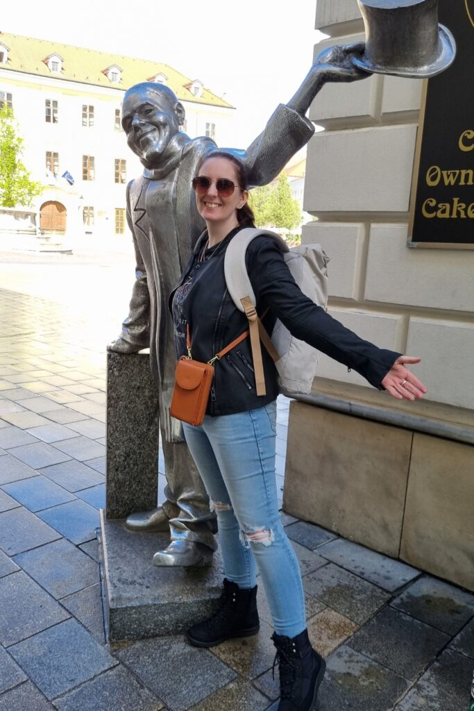 Me with the silver statue of Schöne Náci in Bratislava, a historic local figure depicted tipping his top hat. The woman smiles, mimicking his outstretched pose, with a backpack and crossbody bag.