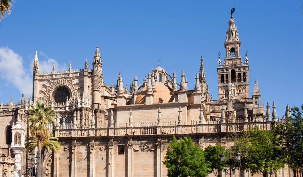 The grand exterior of Seville Cathedral with its intricate Gothic architecture, domes, and spires, including the iconic Giralda tower, all set against a clear blue sky.