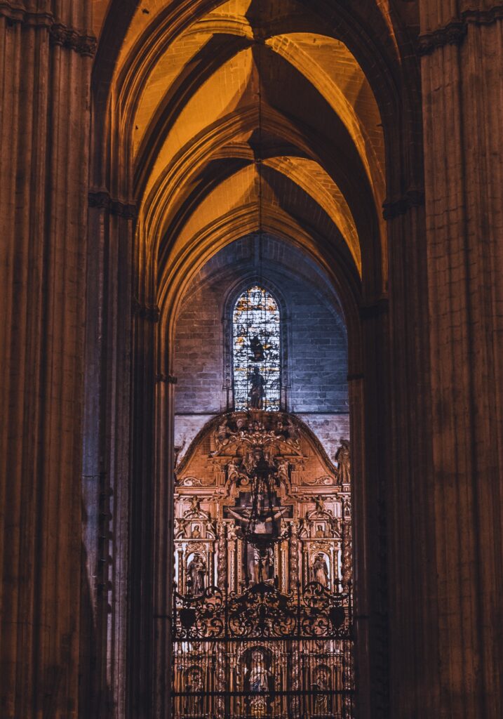 Inside Seville Cathedral, golden light spills over the soaring vaulted arches and a richly detailed altar with religious figures and a stained-glass window above.