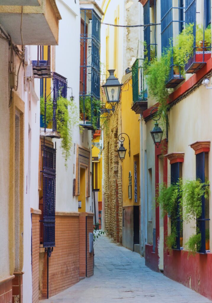 A narrow, colorful alleyway in Seville, Spain, lined with traditional buildings featuring wrought-iron balconies, hanging lanterns, and cascading green plants. A “Restaurante” sign peeks out along the rustic stone wall, inviting passersby into the cozy scene.