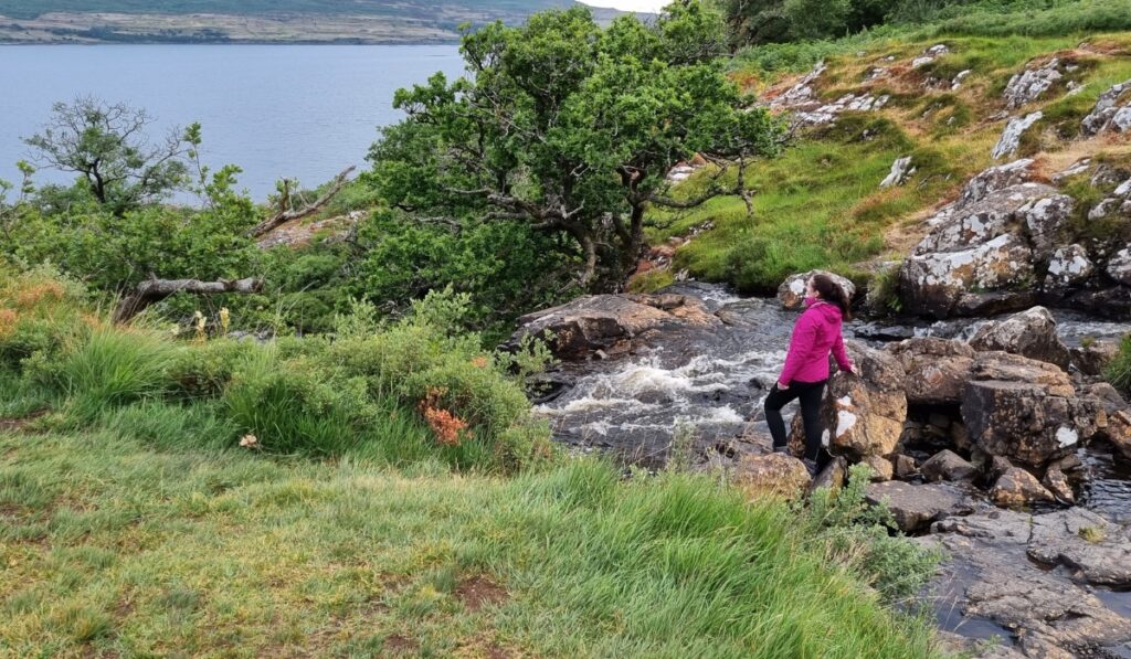 Me wearing my bright pink rain jacket and black pants stands on rocks beside a rushing stream, surrounded by lush greenery and overlooking a peaceful lake. The scene captures a rugged outdoor landscape with dramatic, overcast lighting, perfect for showcasing waterproof hiking gear.
