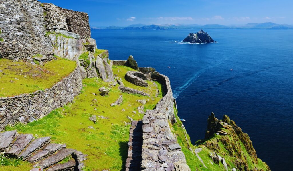 Ancient stone structures and winding paths cling to the steep cliffs of Skellig Michael, an island off the coast of Ireland. The deep blue Atlantic stretches into the distance, with the jagged Little Skellig island visible offshore under a bright blue sky.