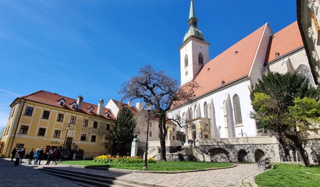 Wide-angle view of St. Martin’s Cathedral in Bratislava, featuring its tall spire, red-tiled roof, and Gothic-style white exterior. The foreground includes a small park with blooming flowers, a statue, and a group of tourists enjoying the sunny day.