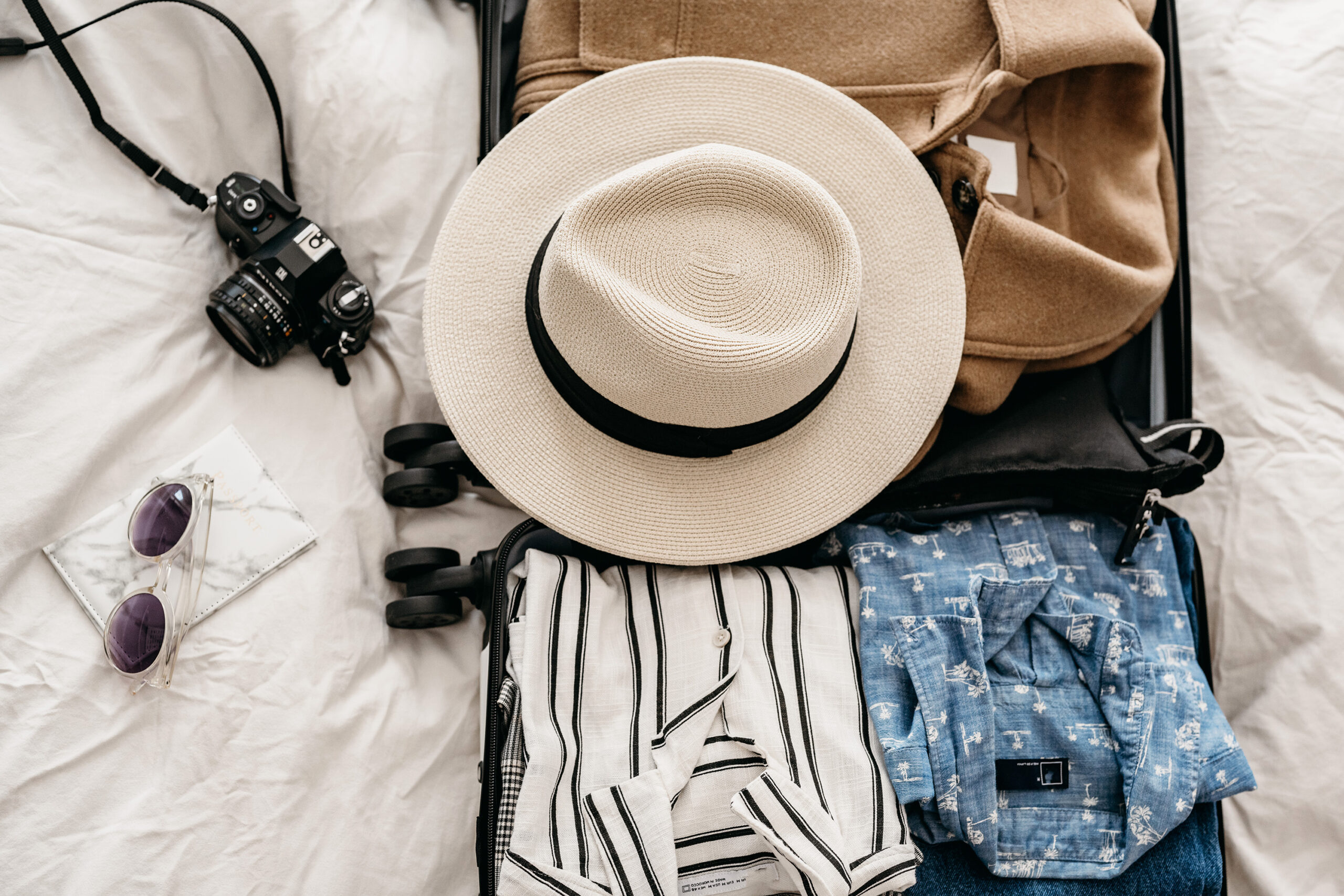 Flat lay of an open suitcase with travel essentials, including a straw hat with a black band, a tan coat, a striped button-up shirt, a blue tropical shirt, and a vintage camera. Nearby on the white bedspread are round sunglasses with a marble case, giving off relaxed vacation vibes.