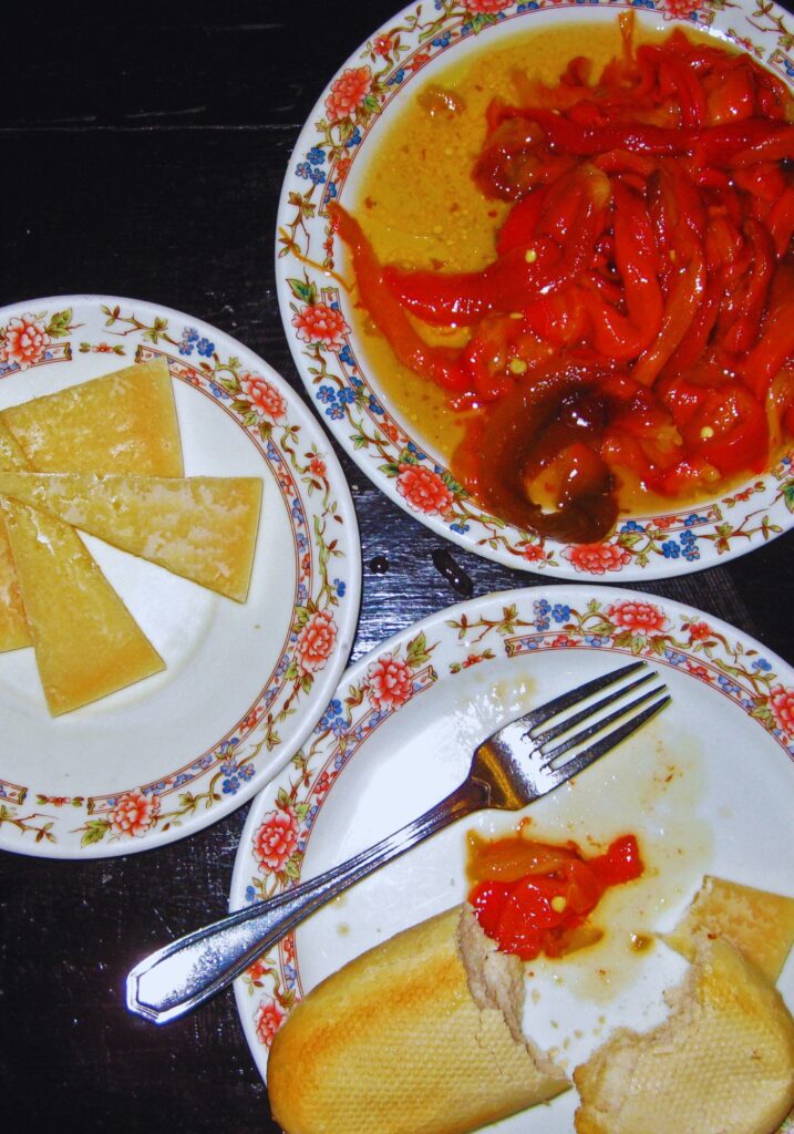 Overhead view of traditional Spanish tapas on floral-patterned plates, featuring slices of Manchego cheese, roasted red peppers in olive oil, and crusty bread on a dark wooden table with a fork.