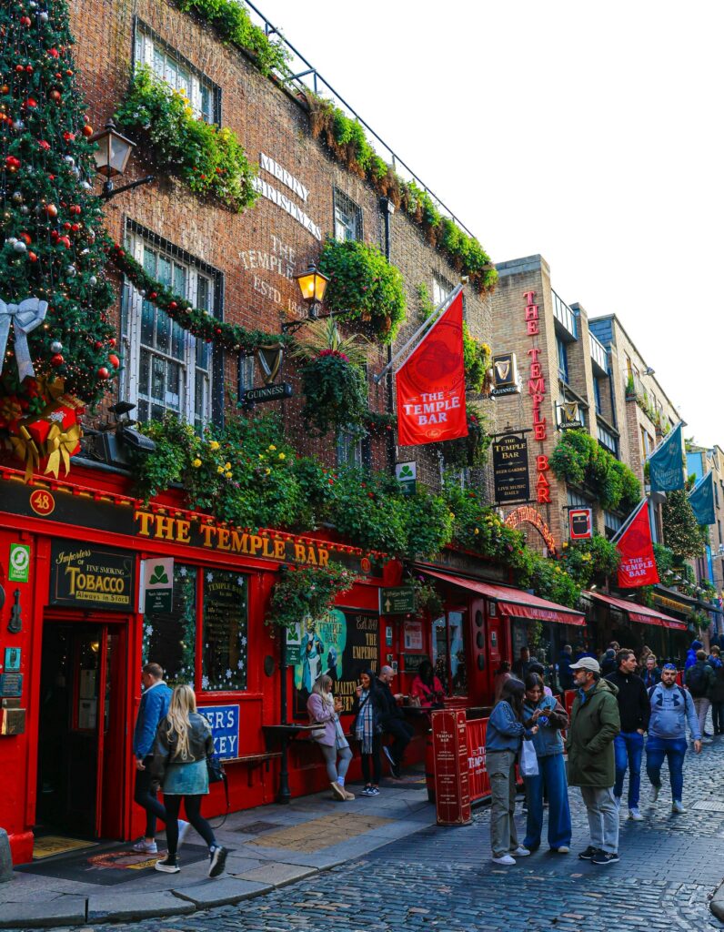 The iconic Temple Bar pub in Dublin, Ireland, is decorated with lush greenery and Christmas lights, with its bright red facade drawing visitors on a cobblestone street. People gather outside taking photos and chatting under the "Merry Christmas" sign painted on the brick wall.