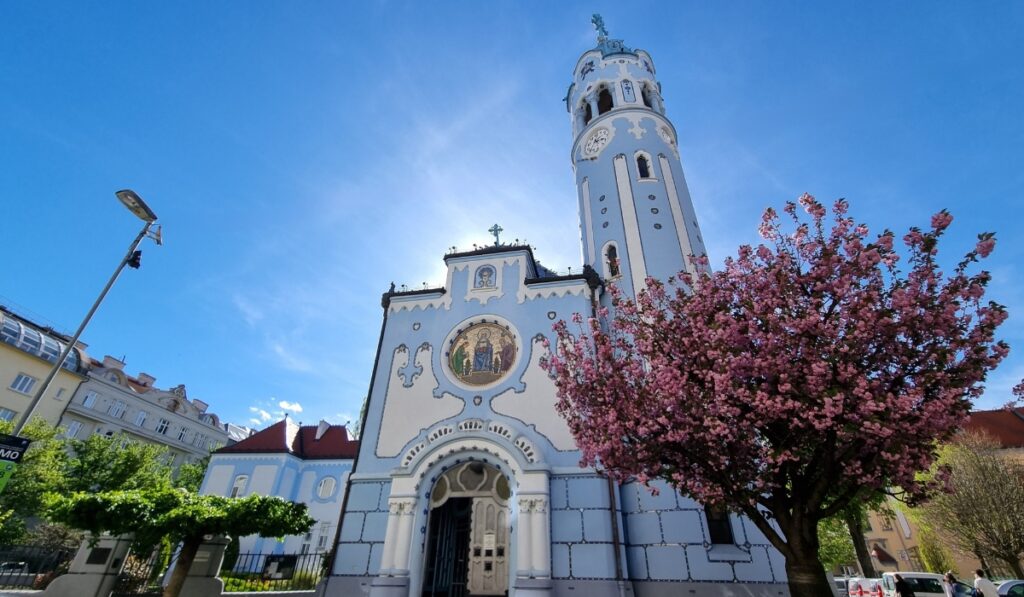 Front view of the Blue Church in Bratislava, showcasing its fairytale-like architecture, mosaic artwork above the entrance, and a blooming cherry blossom tree in the foreground.