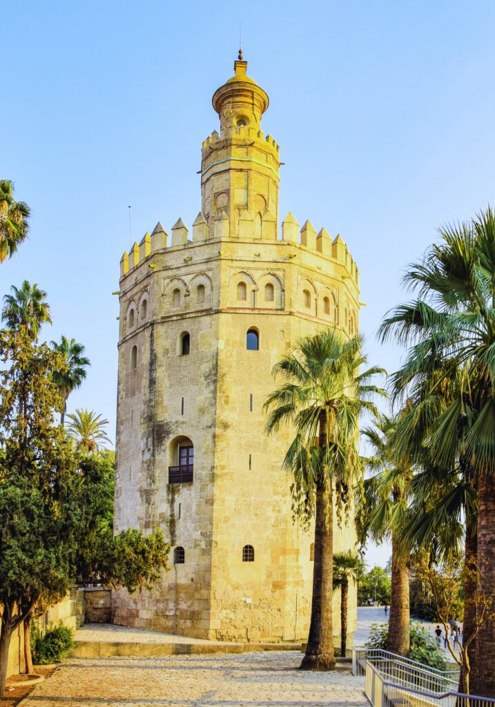 Torre del Oro, a historic 13th-century military watchtower in Seville, Spain, glows in golden sunlight. The dodecagonal tower is surrounded by palm trees and cobblestone paths, set against a clear blue sky.