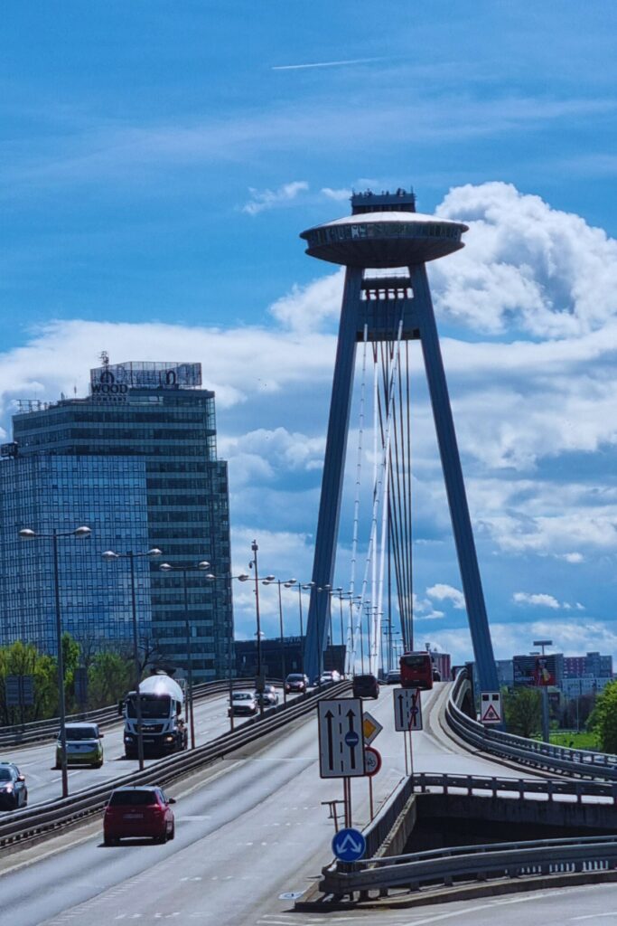 View of Bratislava’s UFO Bridge, officially the SNP Bridge, featuring a futuristic UFO-shaped observation deck atop its tall tower. The bridge spans the Danube River, with traffic flowing beneath a partly cloudy sky and modern high-rises in the background.