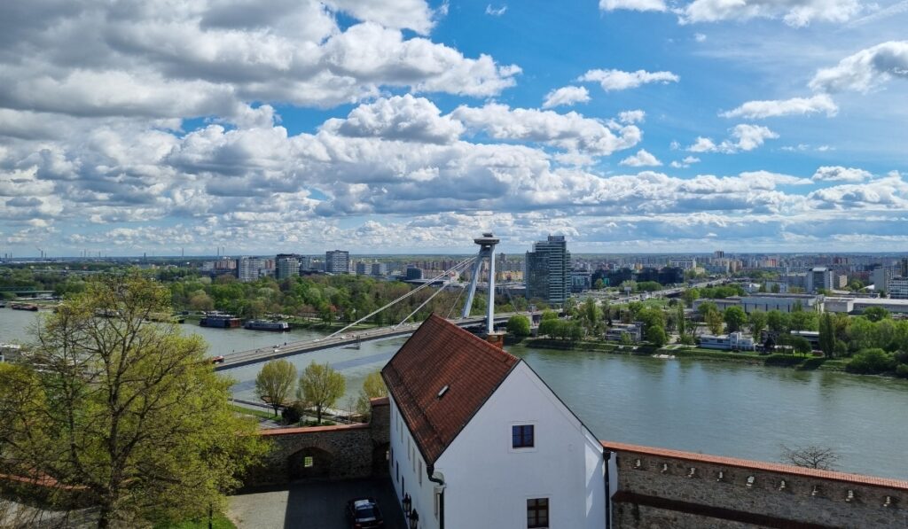 Panoramic view of Bratislava from the castle, featuring the UFO Bridge spanning the Danube River, with the modern cityscape and lush greenery on both sides under a vibrant sky filled with fluffy clouds.