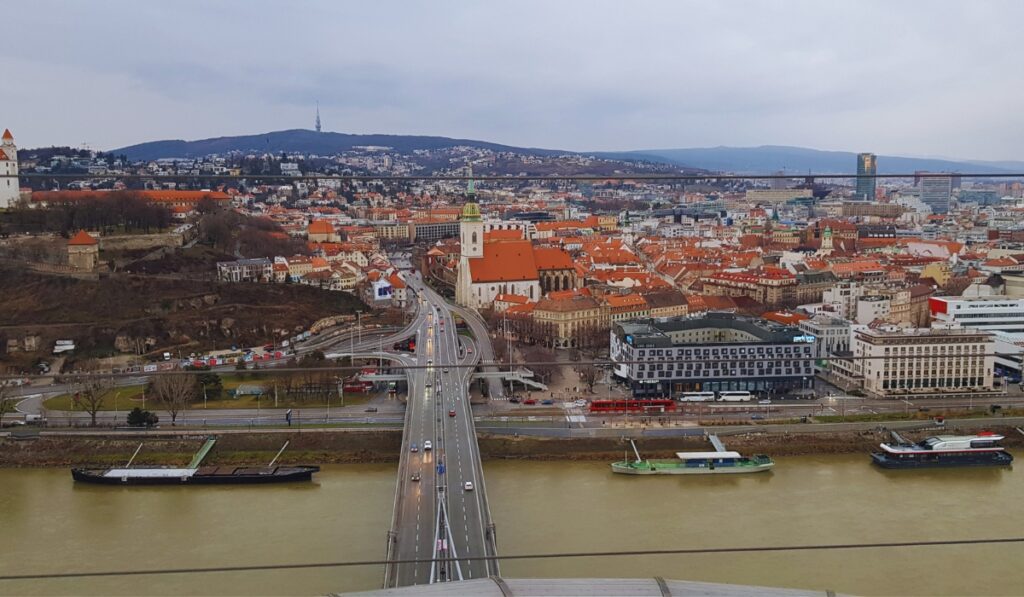 Aerial view of Bratislava from the UFO Tower, overlooking the Danube River, with the SNP Bridge leading into the Old Town. St. Martin’s Cathedral stands out among red-roofed buildings, with Bratislava Castle on a hill to the left and cityscape stretching into the Carpathian hills.