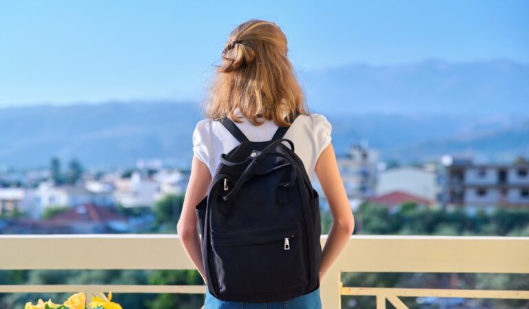 A woman with light brown hair, wearing a white t-shirt and blue jeans, stands on a balcony with a black backpack on her back, looking out at a cityscape with blurred buildings and mountains in the distance under a clear blue sky.