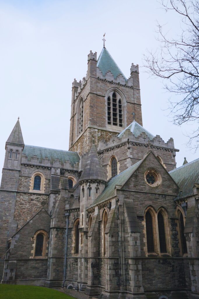 The gothic-style Christ Church Cathedral in Dublin, built of stone with arched windows, pointed towers, and a tall central spire, set against a pale sky.