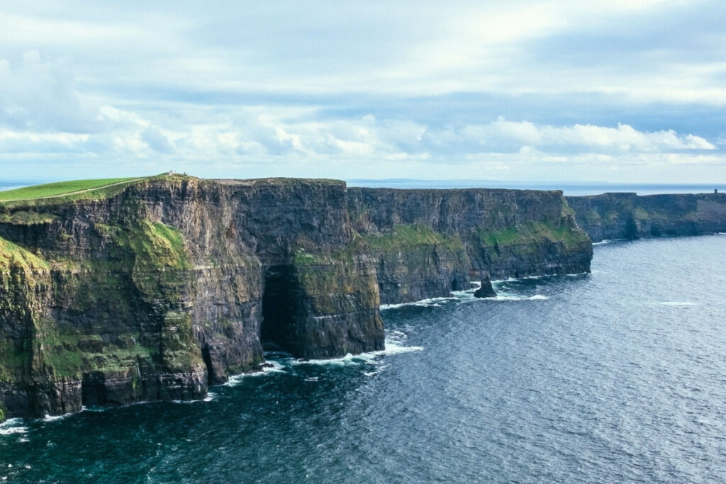 Wide-angle view of the Cliffs of Moher in Ireland, showing dramatic vertical cliffs covered in patches of green grass meeting the choppy Atlantic Ocean under a cloudy sky.