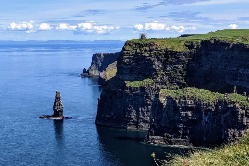 Coastal shot of the Cliffs of Moher on a clear day, with deep blue waters, striking vertical rock formations, and a lone sea stack rising from the ocean.
