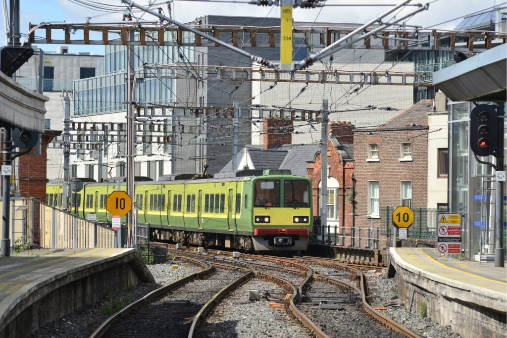 A green DART train travels along curved railway tracks through a cityscape of modern glass buildings and classic brick homes in central Dublin, beneath a web of overhead electric cables.