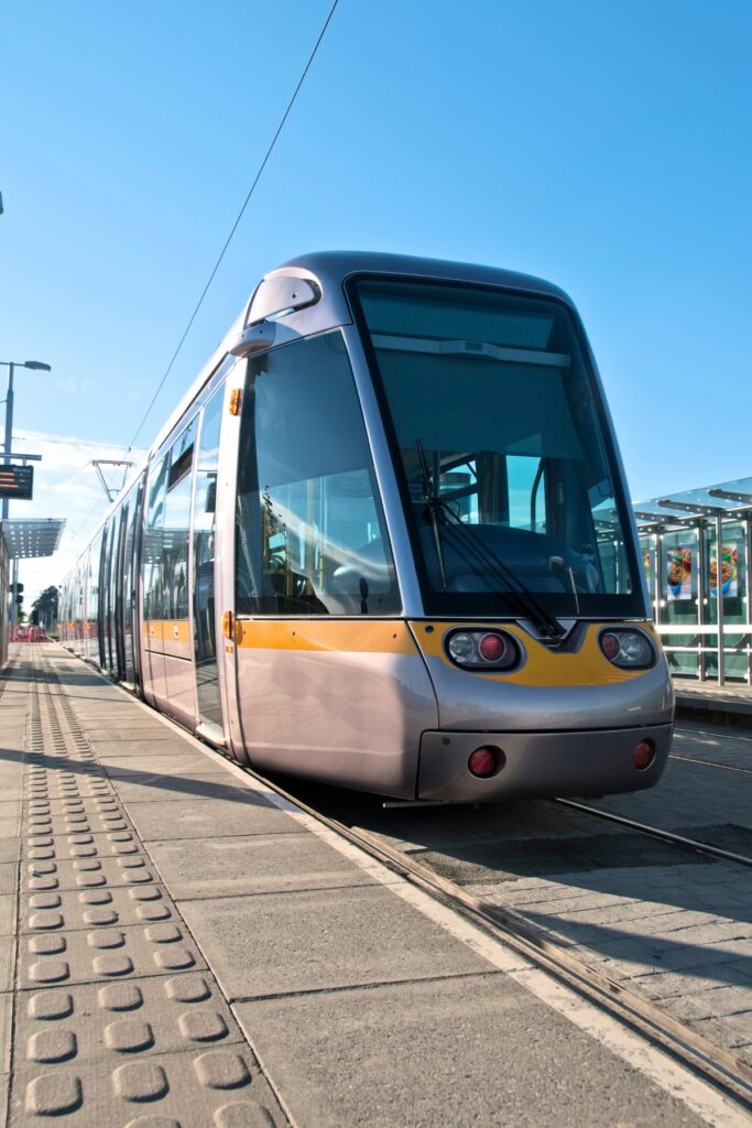 A sleek silver and yellow Luas tram stands at a modern station platform under a bright blue sky, ready to transport passengers across Dublin.