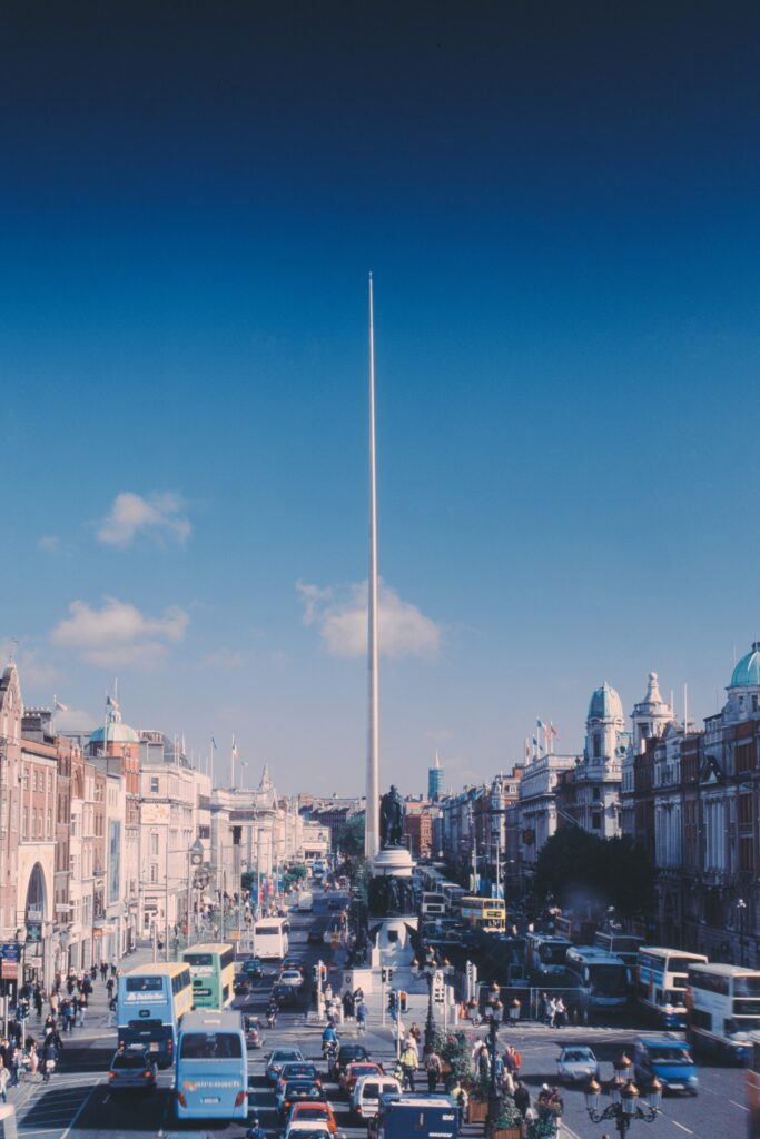 The Spire of Dublin stands tall and slender at the center of busy O'Connell Street, surrounded by historic buildings, statues, and dense city traffic.