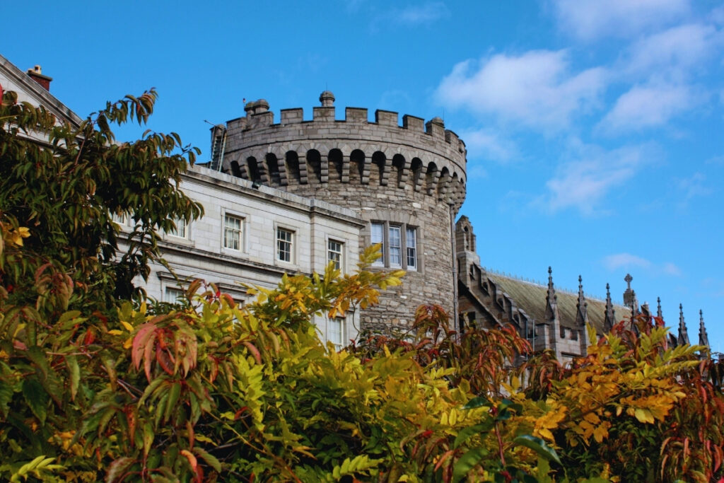 A turreted stone tower of Dublin Castle rises above a mix of autumn-colored foliage and white Georgian architecture, under a bright blue sky.