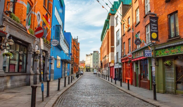Colorful cobblestone street in Dublin’s Temple Bar district, lined with vibrant buildings, including the bright blue Bad Bobs Temple Bar and the red Stage Door Café advertising "Dublin’s Best Breakfast." Irish flags and festive bunting hang above the quiet pedestrian street on a clear day.