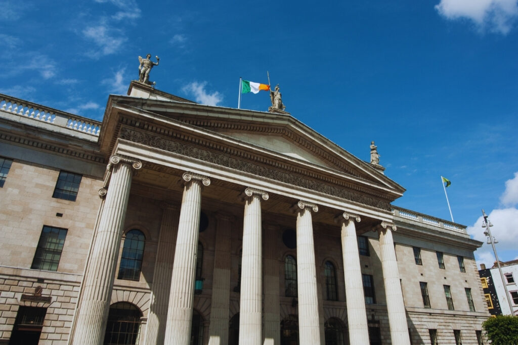 The General Post Office in Dublin, a neoclassical building with grand columns and an Irish flag flying above, set against a blue sky with light clouds.