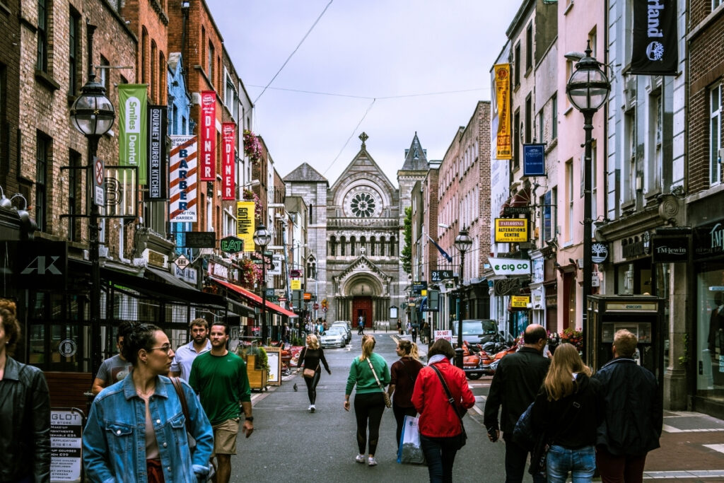 Crowds of people walk along vibrant Grafton Street in Dublin, lined with shops, eateries, and colorful signage, with the stone façade of St. Teresa’s Church visible in the distance.