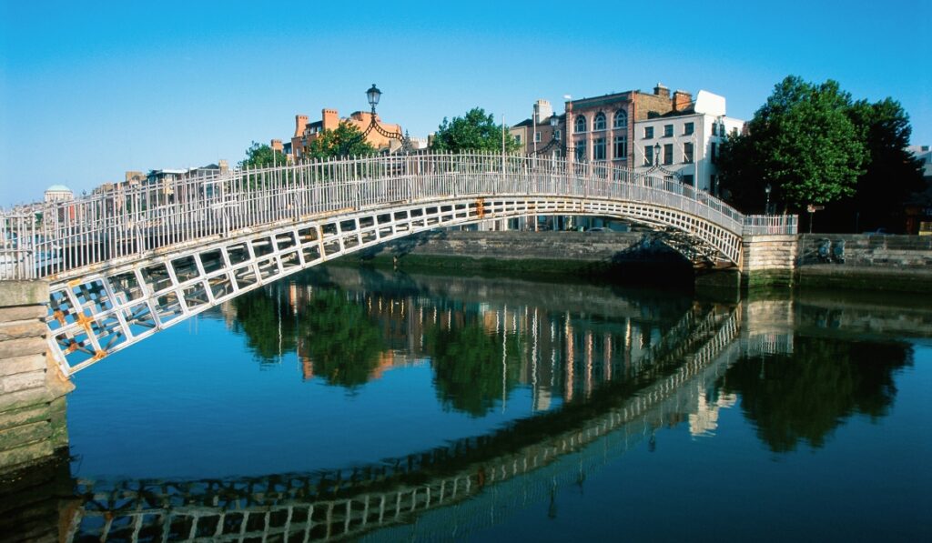 The Ha'penny Bridge arches gracefully over the calm River Liffey in Dublin, with its white iron structure reflecting in the water under a clear blue sky, surrounded by historic brick buildings and greenery.