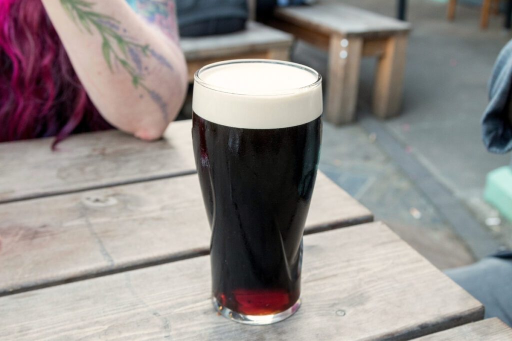 A freshly poured pint of Guinness with a creamy white head sits on a rustic wooden table at an outdoor pub, with a tattooed arm and pink hair partially visible in the background.