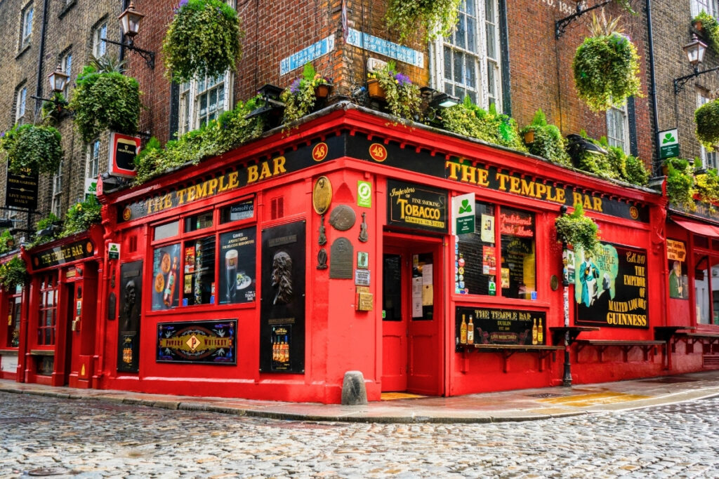 The famous red façade of The Temple Bar pub in Dublin, adorned with hanging plants and classic signage, sits on a cobblestone corner known for live music and Irish whiskey.