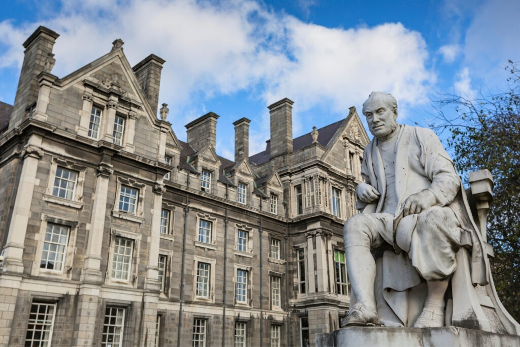 A marble statue of a seated man in front of the ornate stone facade of Trinity College Dublin, with chimneys, tall windows, and blue sky overhead.