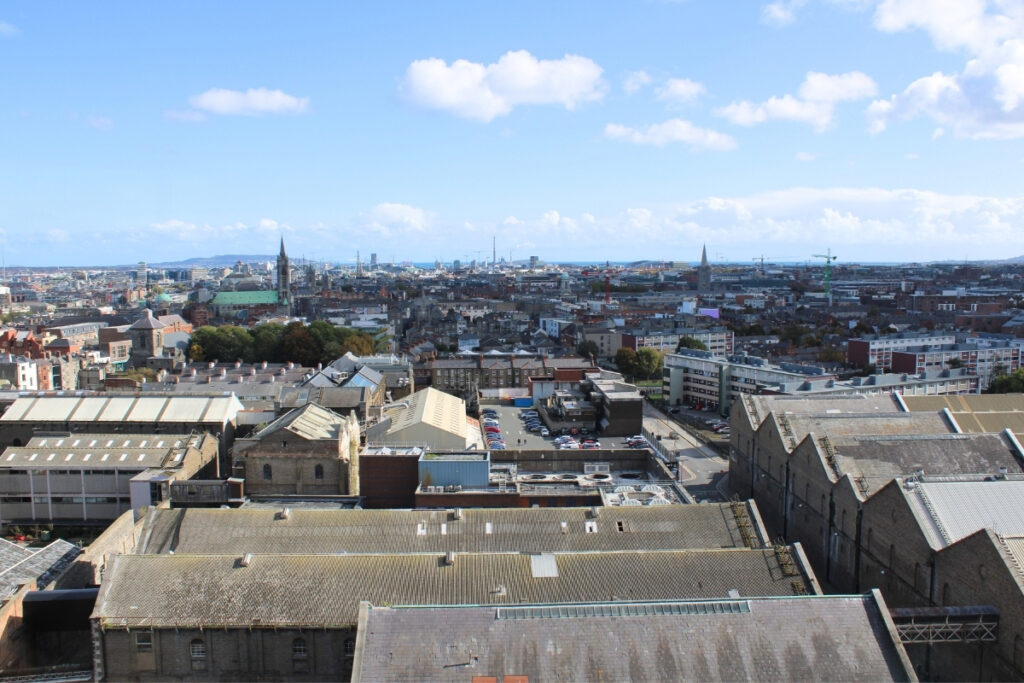 Panoramic view of Dublin city from the Gravity Bar at the Guinness Storehouse, showing a mix of industrial rooftops, church spires, and modern buildings stretching toward the Irish Sea under a bright blue sky.
