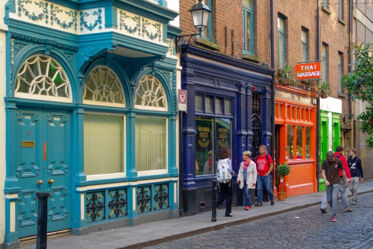 A lively Dublin street scene featuring a row of vibrantly colored storefronts, including a teal Georgian-style building, a deep navy shop labeled "House of Horrors," and a bright orange Thai massage parlor. Several pedestrians in casual attire walk and chat along the cobblestone sidewalk, adding to the energetic, urban vibe.