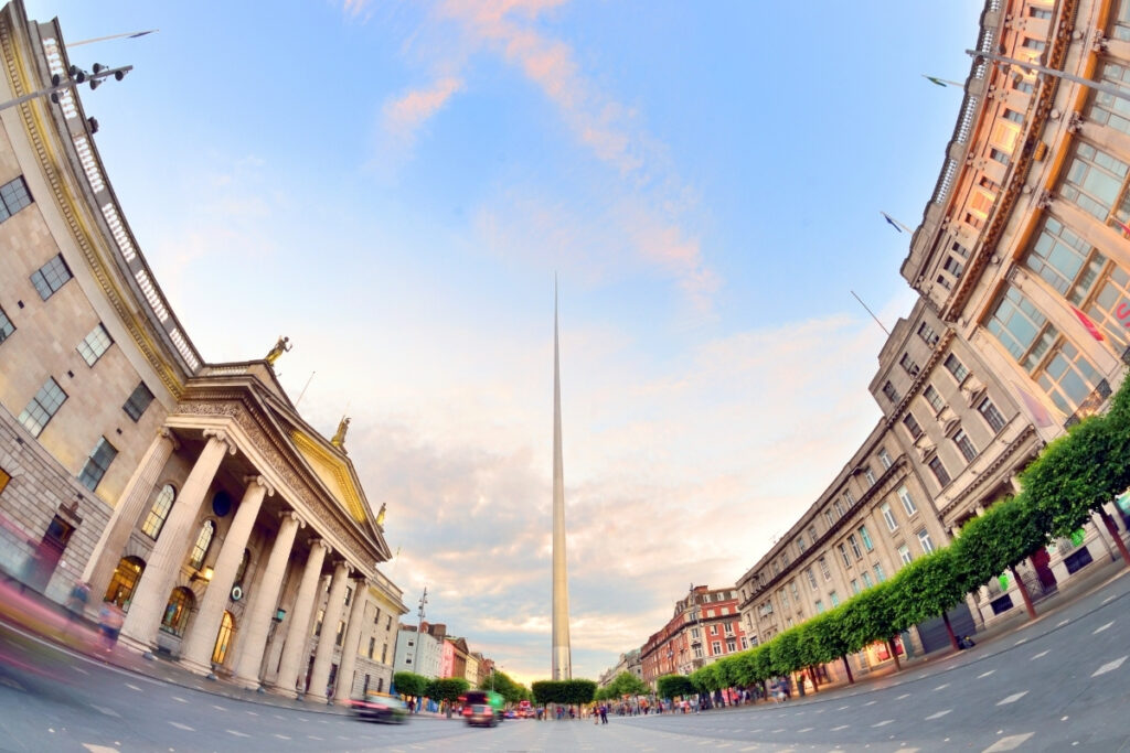 Wide-angle view of O’Connell Street in Dublin at sunset, featuring the Spire of Dublin towering in the center, flanked by classical buildings on both sides, including the General Post Office (GPO) with its grand columns and statues. The street is slightly blurred with motion from cars and people, creating a dynamic, lively city atmosphere.