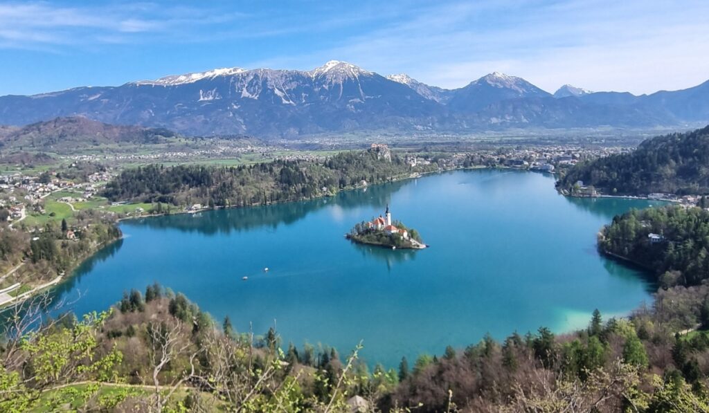 This image shows the picturesque Lake Bled in Slovenia, featuring the iconic Bled Island with the Church of the Assumption rising from its center. The emerald-blue water, surrounding forested hills, and distant mountains create a tranquil and scenic landscape.