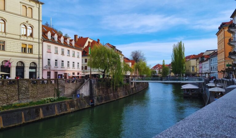 A colorful riverside scene in Ljubljana, Slovenia, showing historic buildings with red-tiled roofs lining the Ljubljanica River. People sit at outdoor cafés under trees and awnings while a modern pedestrian bridge crosses the calm water in the background.