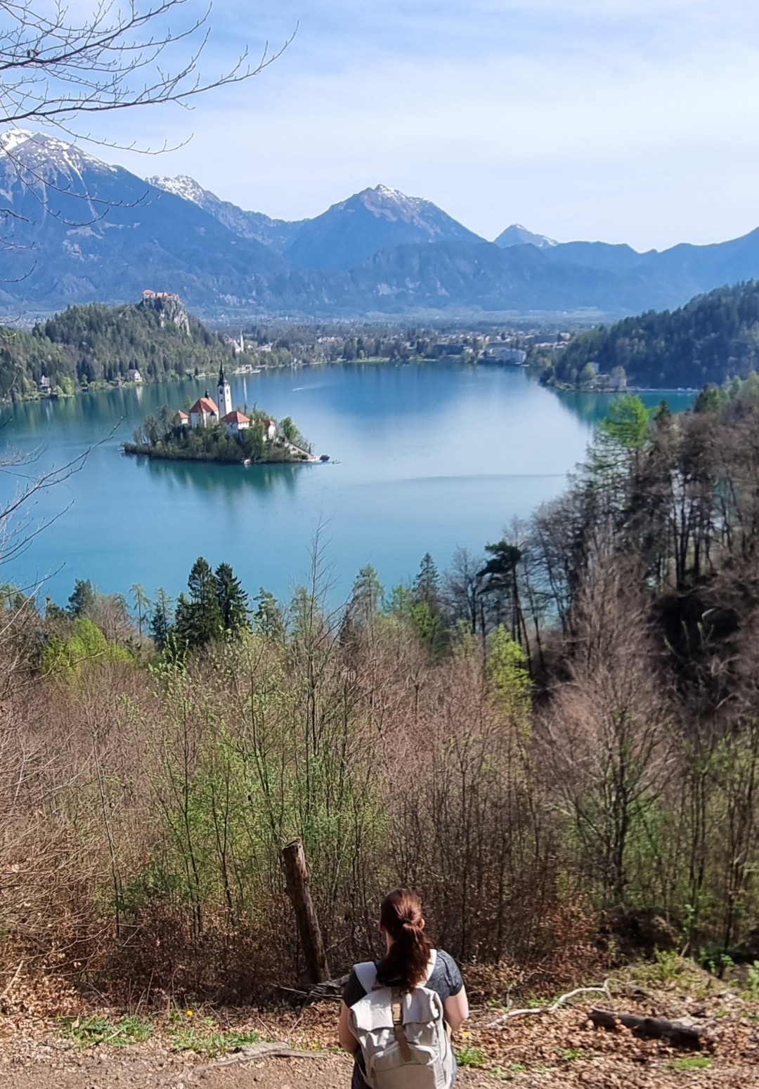Me, sitting on a rock at a scenic viewpoint overlooking Lake Bled and its island with the Church of the Assumption, surrounded by calm blue water and framed by forested hills and snow-capped mountains. The image captures a peaceful moment of nature and travel in Slovenia.